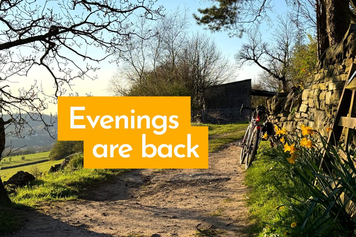 Text 'Evenings are back' on photo of bicycle propped against stone wall on dirt track above valley. Evening sun illuminates daffodils.